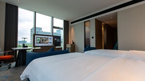 Habitación de hotel con una cama grande, un escritorio con computadora, televisión y vista a la ciudad a través de amplias ventanas.