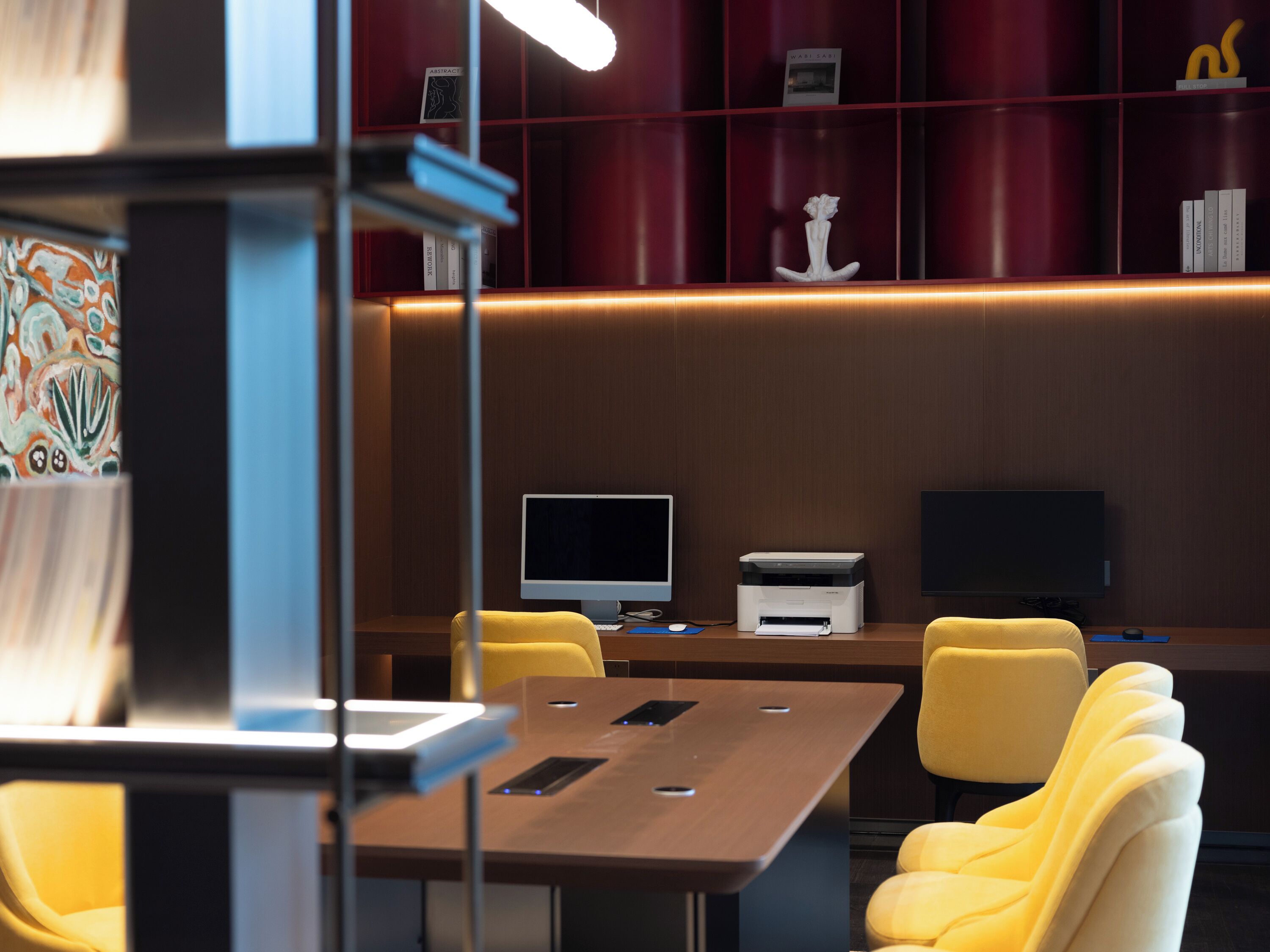 A modern office space with a wooden desk, two yellow chairs, a computer monitor, a printer, and a bookshelf with various items.