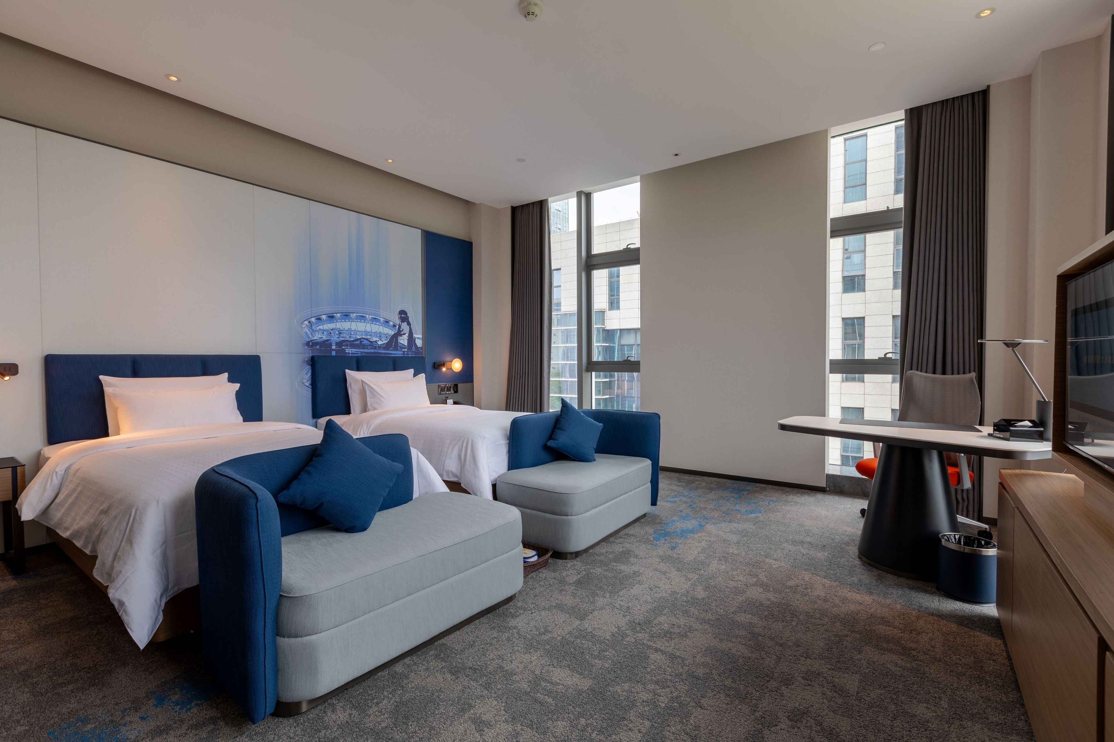Une chambre d’hôtel moderne équipée de deux lits, d’un canapé, d’un bureau et d’une grande fenêtre donnant sur la ville.