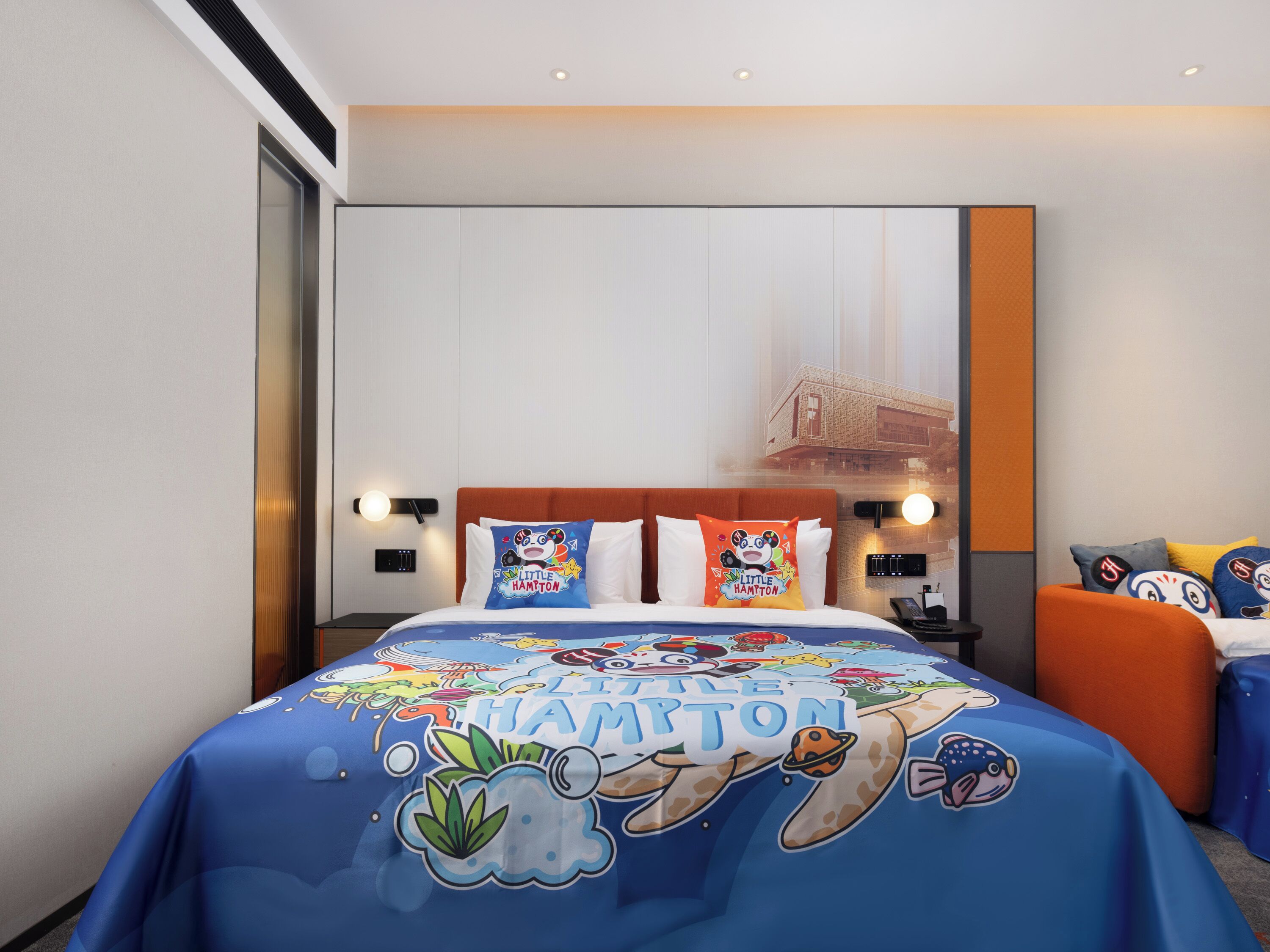 Una habitación de hotel con una cama que presenta un cubrecama azul con el texto 'Little Hampton' e ilustraciones, un cabecero rojo y vistas de un puente a través de una ventana.