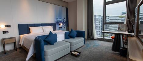 A modern hotel room with a large bed, a sofa, a desk, and a view of the cityscape through large windows.