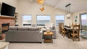 A spacious living room with a fireplace, a large sofa, and a dining area with wooden chairs. - Unreal Panoramic Oceanfront and Beach Views, Hot Tub, New Game Room, Fire-table, 1/2 Mile to Town (Rockaway Beach)