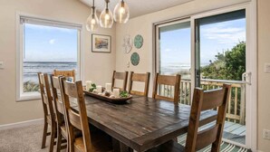 A dining area with a wooden table, six chairs, and a view of the ocean through large sliding glass doors. - Unreal Panoramic Oceanfront and Beach Views, Hot Tub, New Game Room, Fire-table, 1/2 Mile to Town (Rockaway Beach)