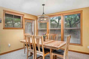 A dining area with a wooden table and chairs, large windows with blinds, and a hanging light fixture.