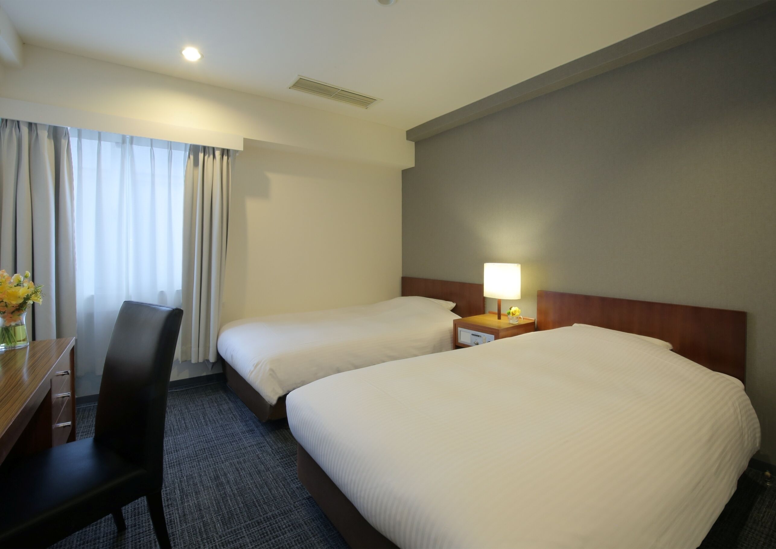 A hotel room with two beds, a desk, a chair, a nightstand with a lamp, and a window with curtains.