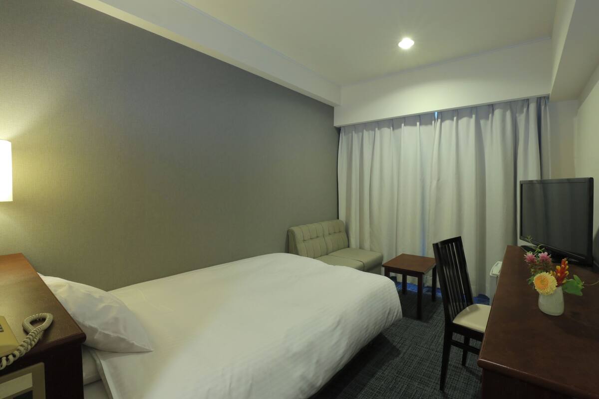 A hotel room with a single bed, a desk with a telephone, a small table, a chair, a TV, and a vase with flowers.