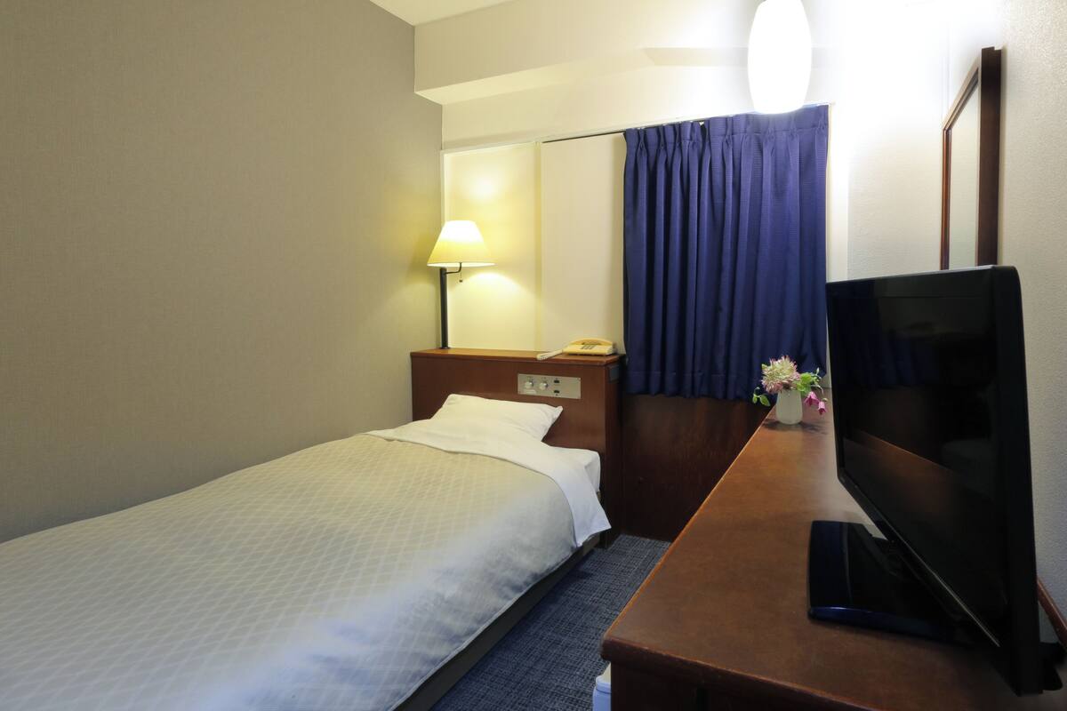 A hotel room with a single bed, a desk with a television, a lamp, and a telephone.