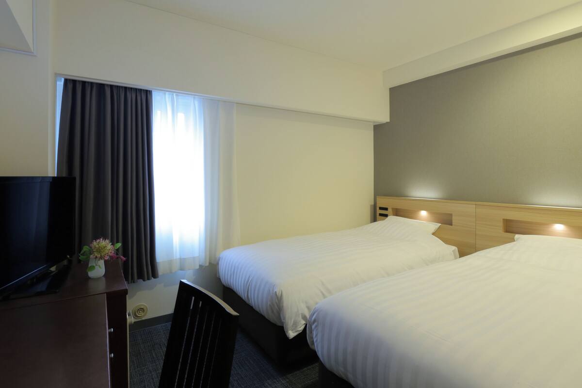 A hotel room with two beds, a desk, a television, and a window with curtains.