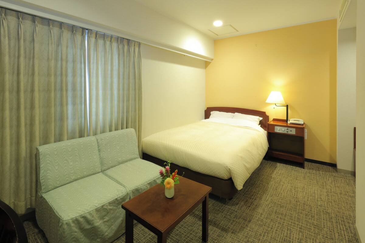 A hotel room with a bed, a sofa, a nightstand with a lamp, and a small table with a vase of flowers.