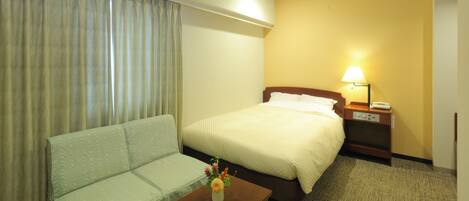 A hotel room with a bed, a sofa, a nightstand with a lamp, and a small table with a vase of flowers.