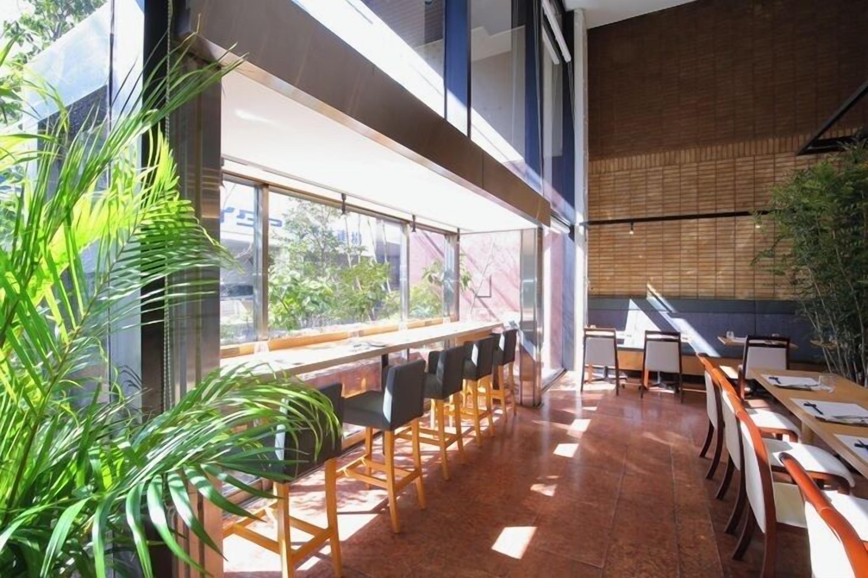 A modern restaurant with long communal tables and chairs, large windows offering views of the outdoors, and a mix of natural and artificial lighting.