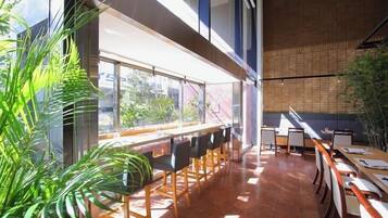 A modern restaurant with long communal tables and chairs, large windows offering views of the outdoors, and a mix of natural and artificial lighting.