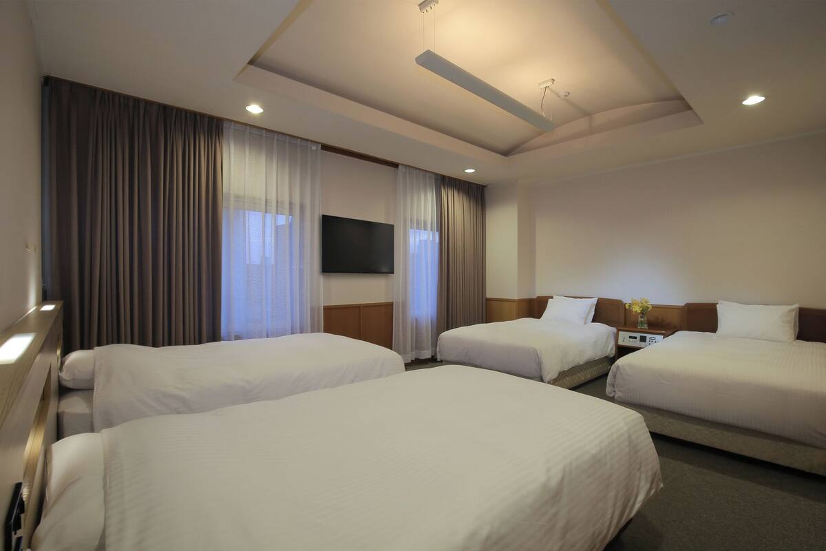 A hotel room with three beds, a flat-screen TV, a desk with a lamp, and a vase with flowers.