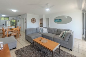 A living room with a grey sofa, a wooden coffee table, a dining table with chairs, and a kitchen area in the background.