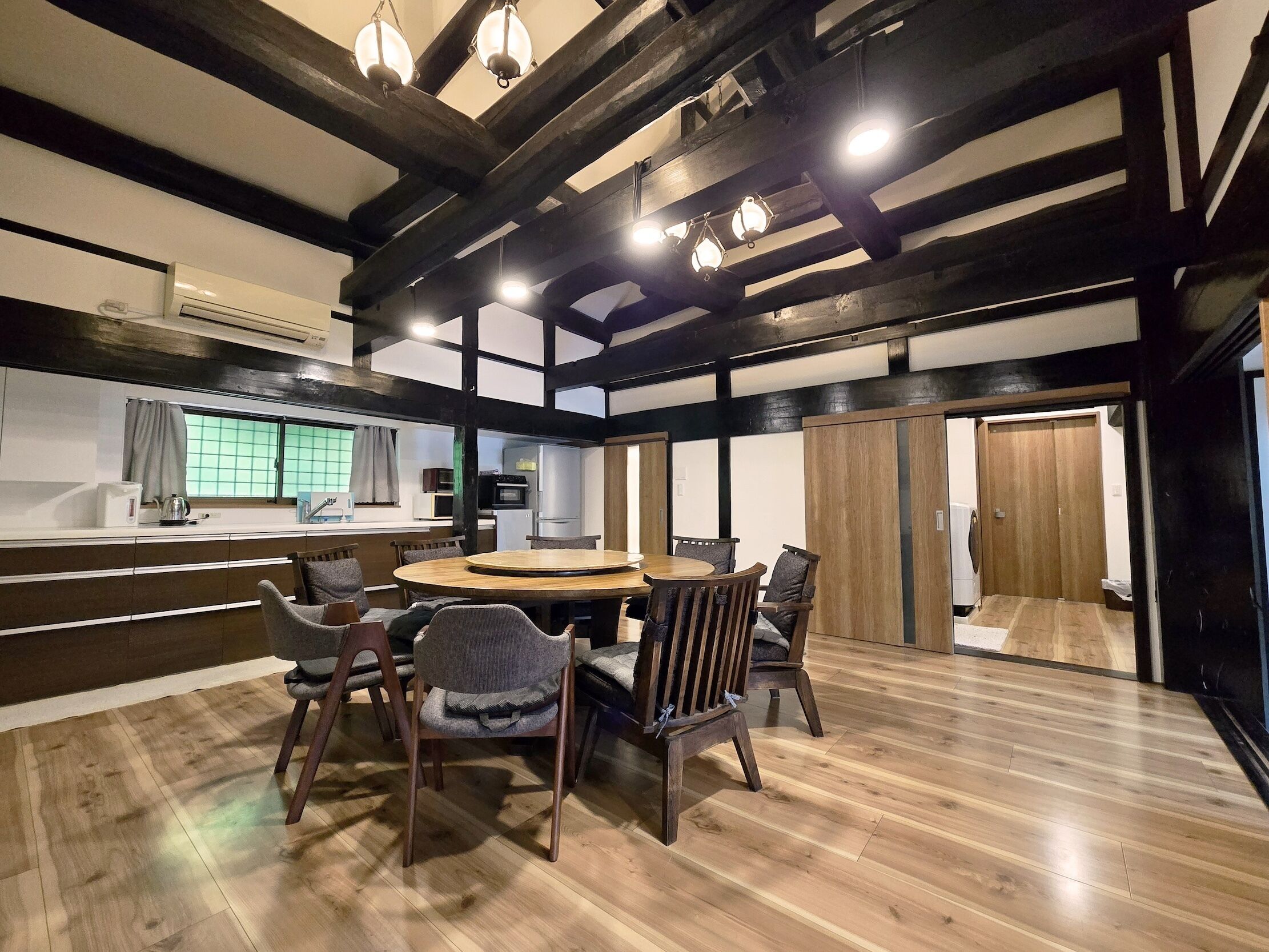 Uma área de jantar com uma mesa de madeira redonda e cadeiras, uma cozinha ao fundo, e estilo arquitetônico japonês tradicional com vigas aparentes e esteiras de tatame.