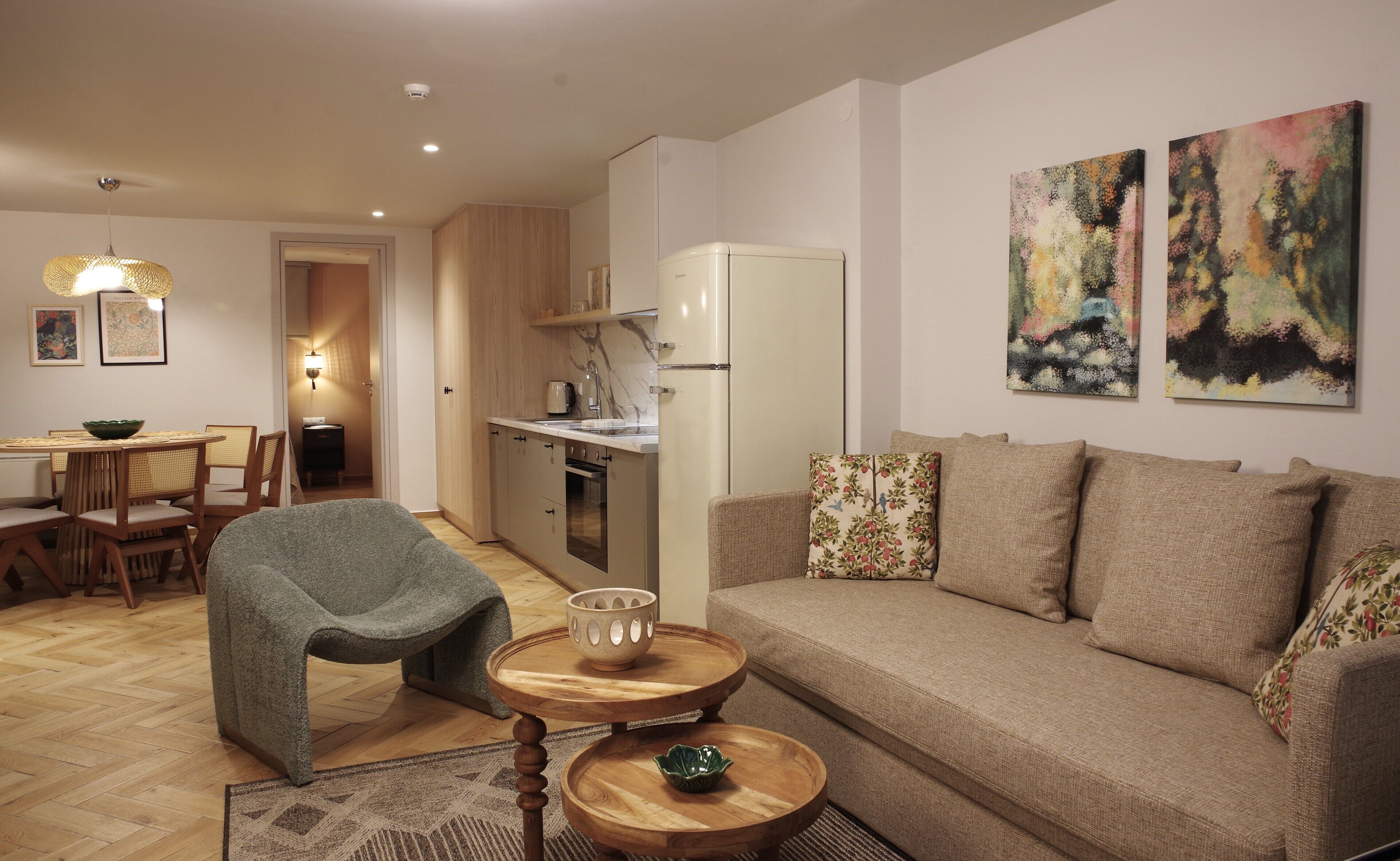 A modern living room with a sofa, a dining table with chairs, a kitchen area with a refrigerator, and artwork on the walls.