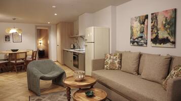 A modern living room with a sofa, a dining table with chairs, a kitchen area with a refrigerator, and artwork on the walls.