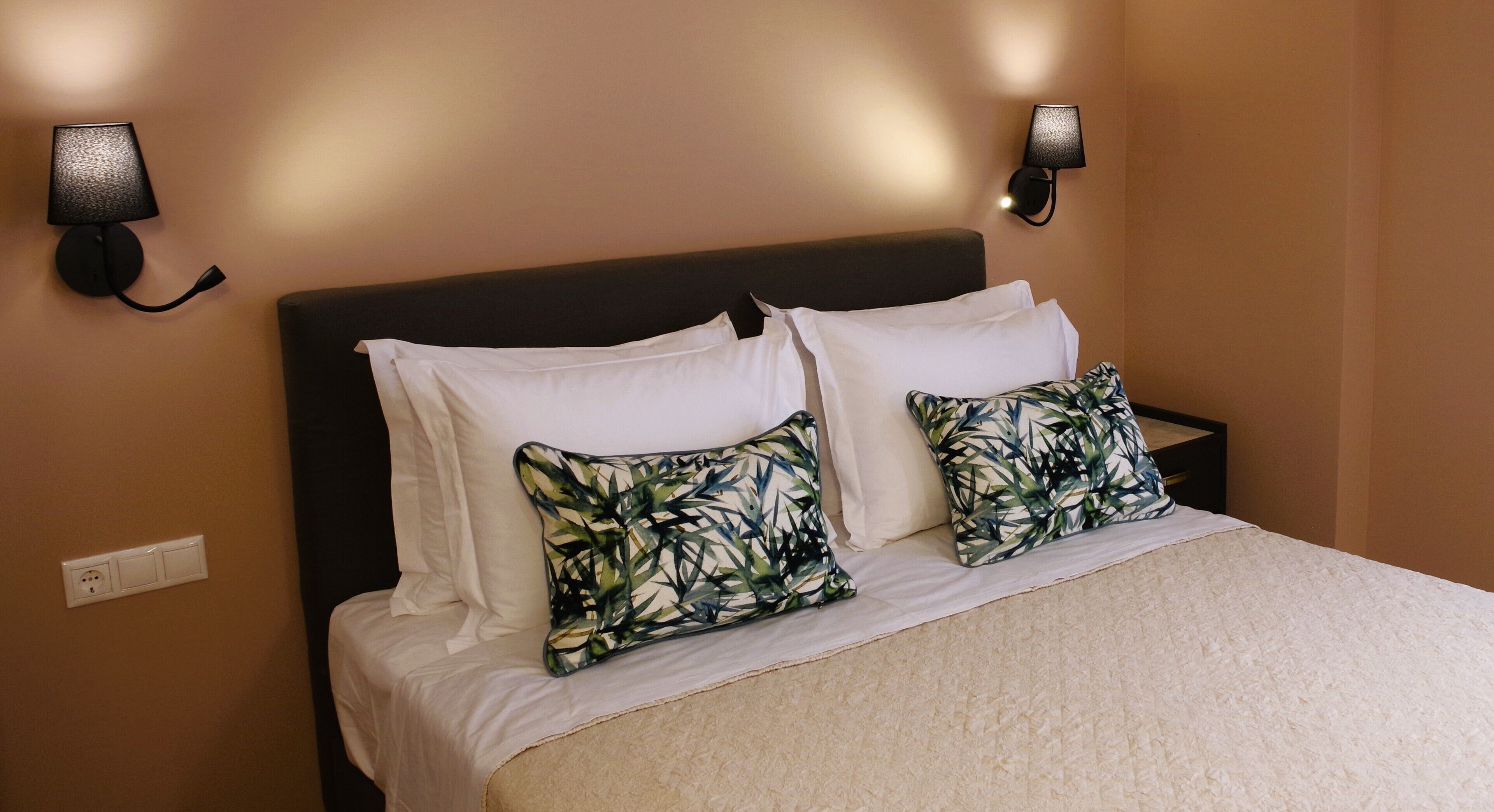 A neatly made bed with white linens and two decorative pillows, a dark headboard, and two wall-mounted lamps on either side.