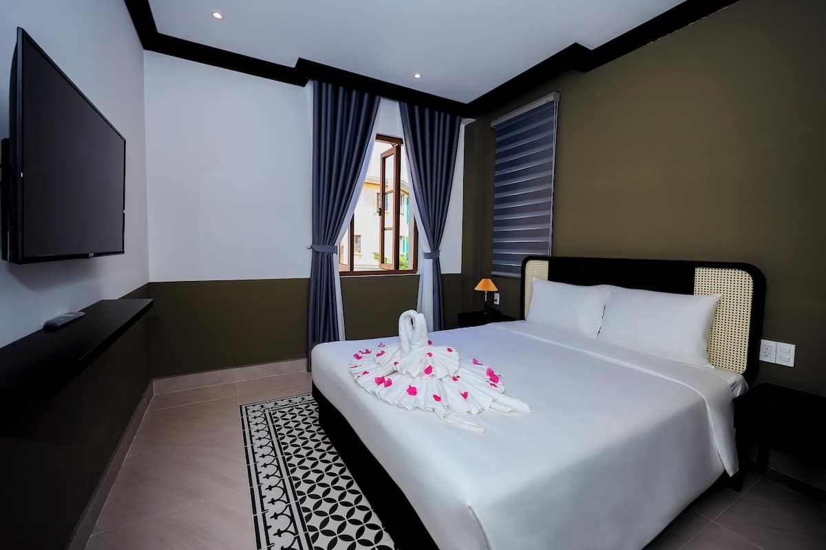A hotel room with a large bed, a flat-screen TV, a window with curtains, and a bedside table with a lamp.