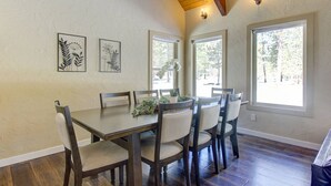 A dining area with a wooden table and chairs, framed botanical artwork on the wall, and large windows with a view of trees.