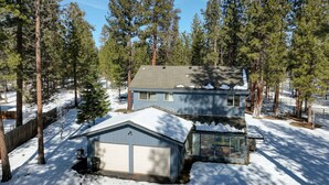 A two-story house with a garage, surrounded by tall pine trees and covered in snow.