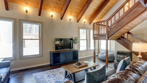 A spacious living room with a wooden ceiling, large windows, a television, a sofa, a coffee table, and a staircase.