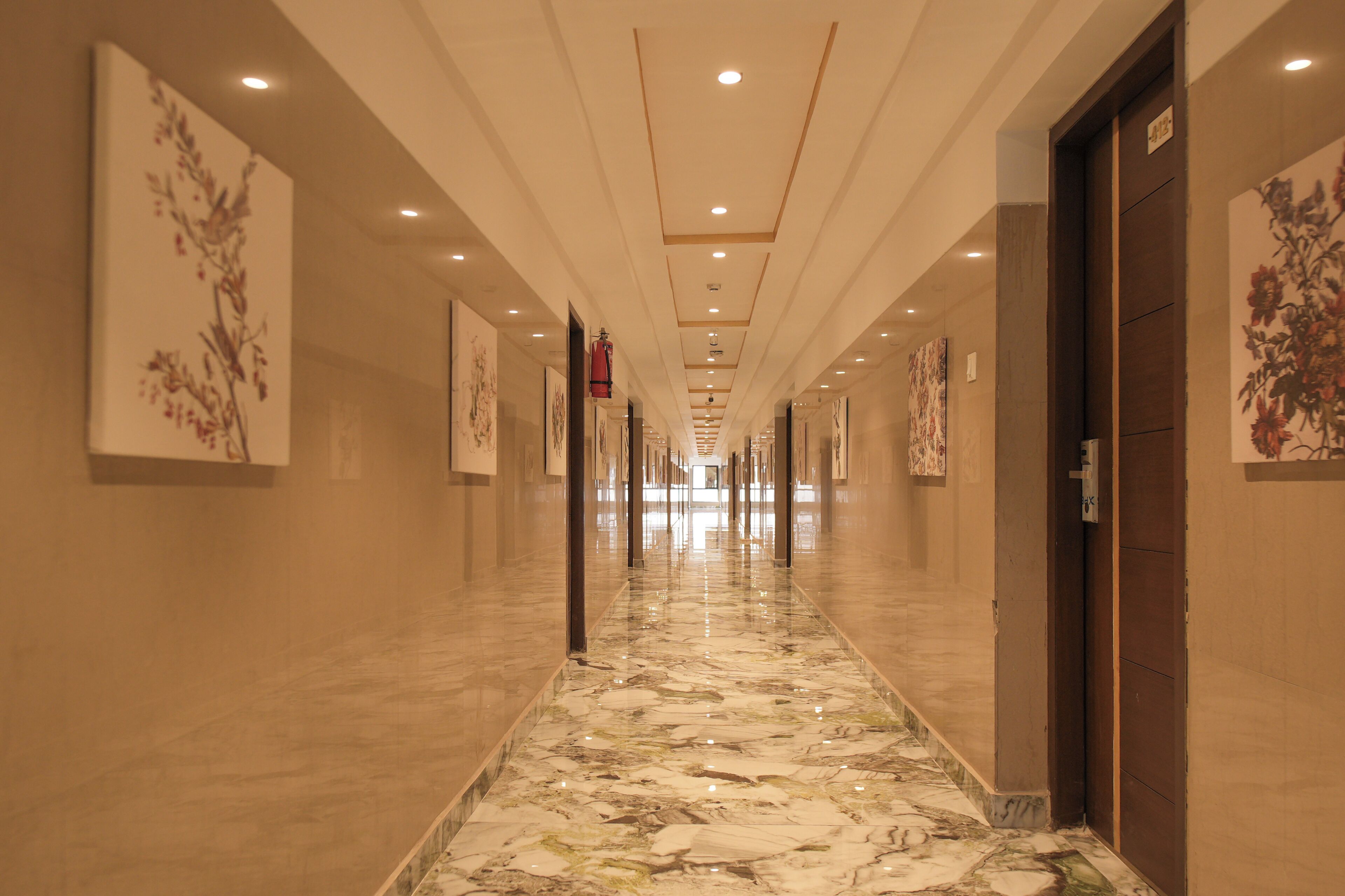 A long, well-lit hallway with marble flooring, flanked by walls with floral artwork, leading to a door on the right.