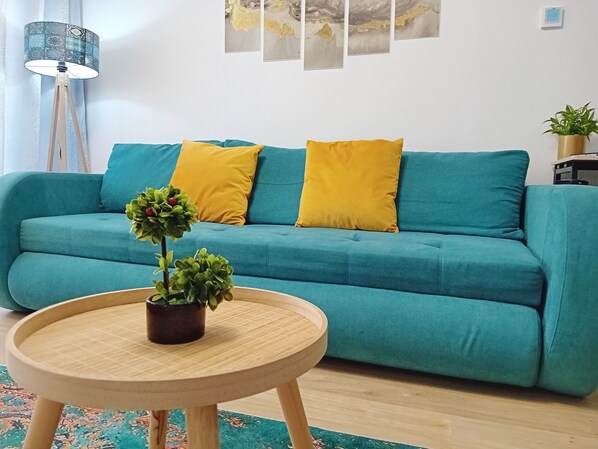 A teal sofa with two yellow cushions, a small wooden table with a potted plant, and a floor lamp.