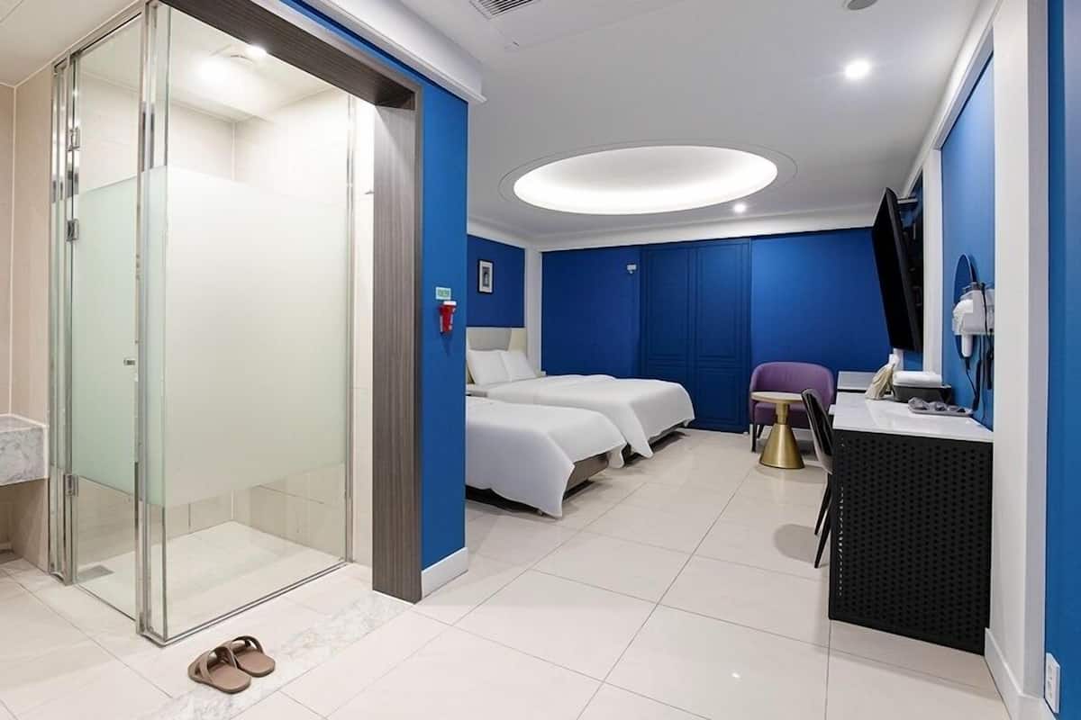 A hotel room with a glass shower enclosure, two beds, a desk with a chair, and a small round table.