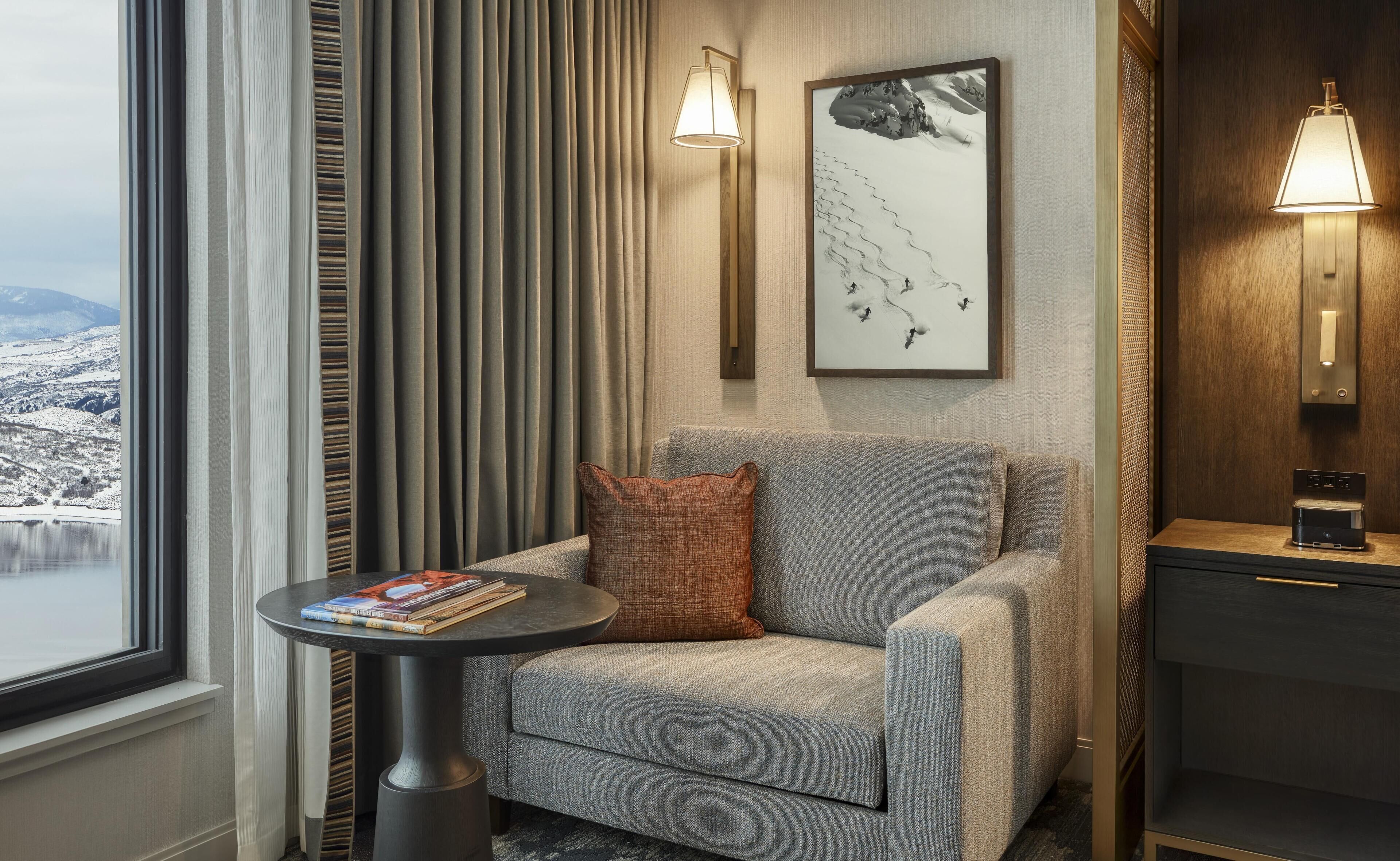 Une chambre d’hôtel avec un fauteuil gris, une table ronde sur laquelle sont posés des livres, un tableau abstrait encadré et une vue sur un paysage enneigé par la fenêtre.