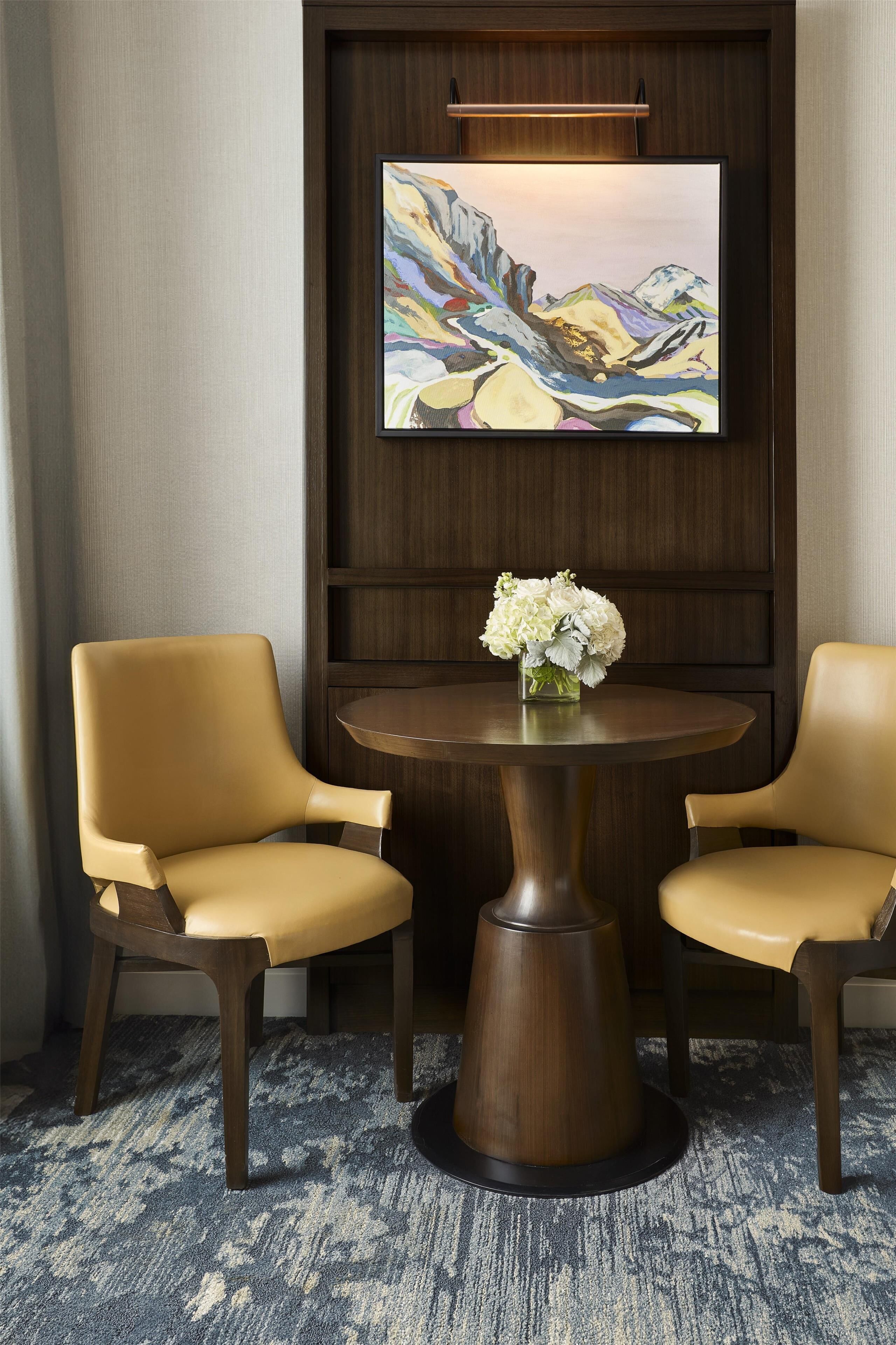 A room with two leather chairs, a round table with a vase of flowers, and a painting on the wall.
