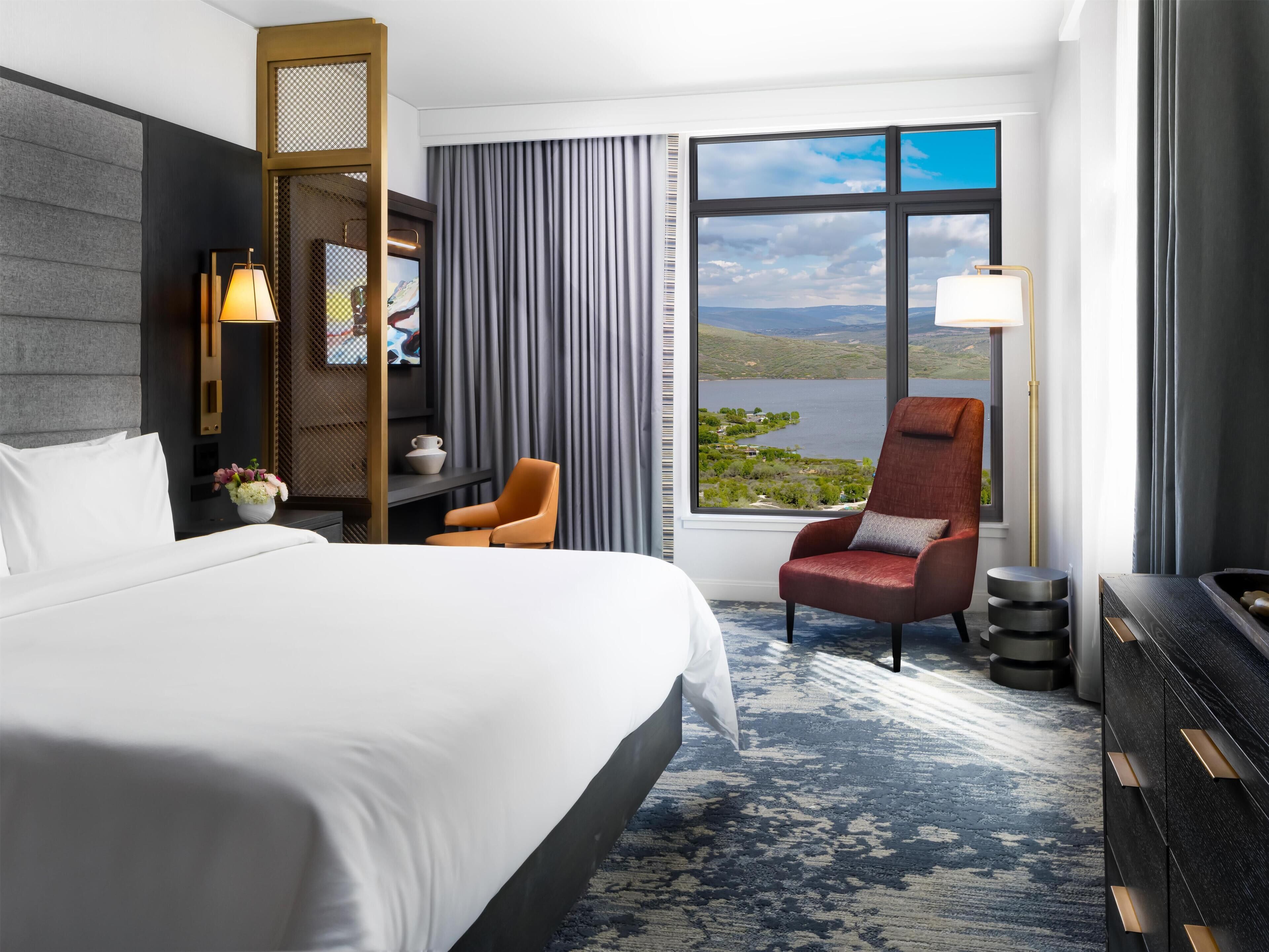 Ein Hotelzimmer mit einem großen Bett, einem Schreibtisch mit Stuhl, Blick auf einen See und moderner Einrichtung.