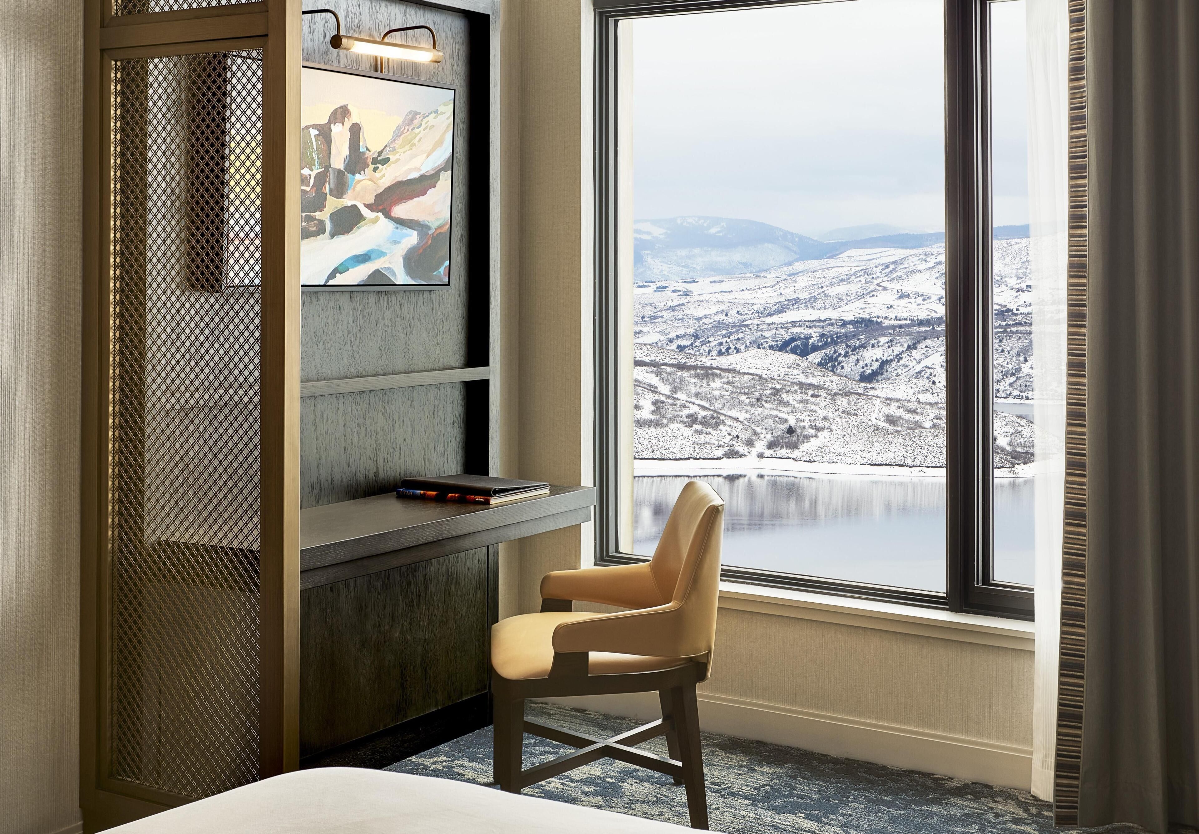 A room with a large window, a chair, a desk with a computer, and a painting.