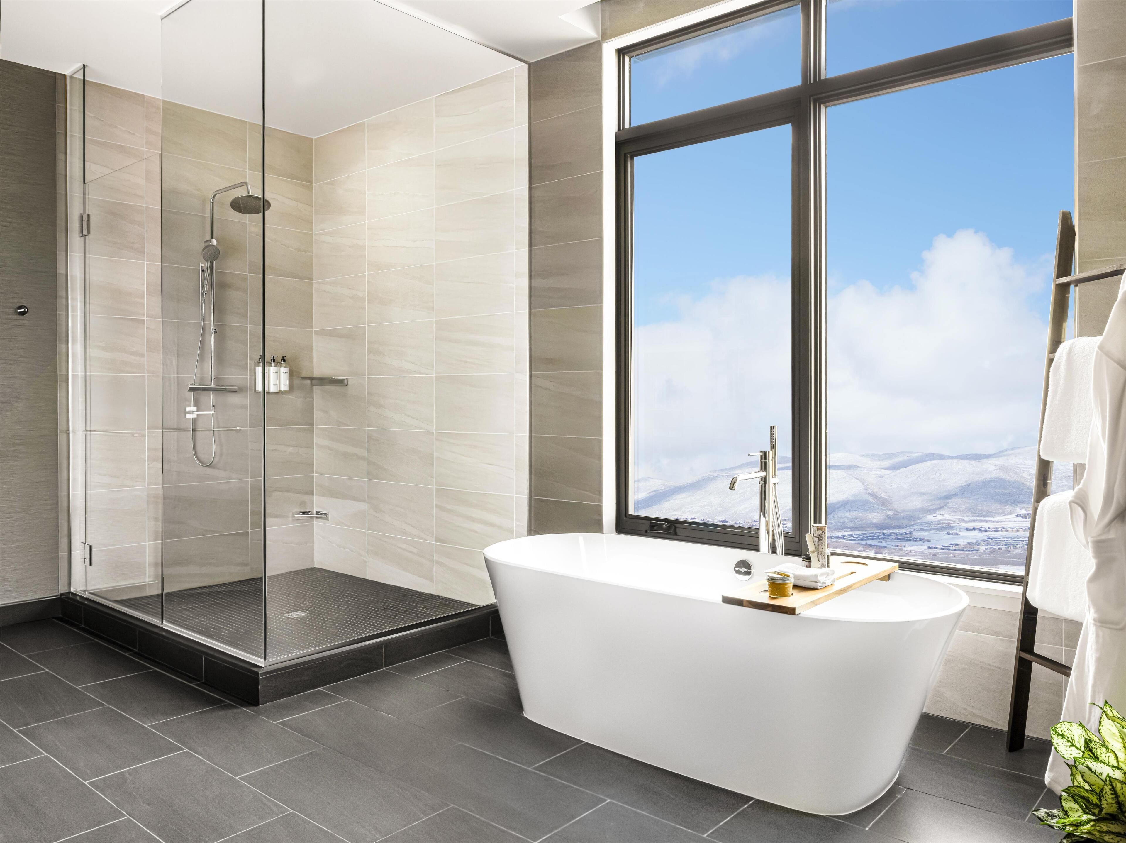 Une salle de bain moderne dotée d’une douche à parois de verre, d’une baignoire sur pieds et d’une grande fenêtre offrant une vue sur des montagnes enneigées.
