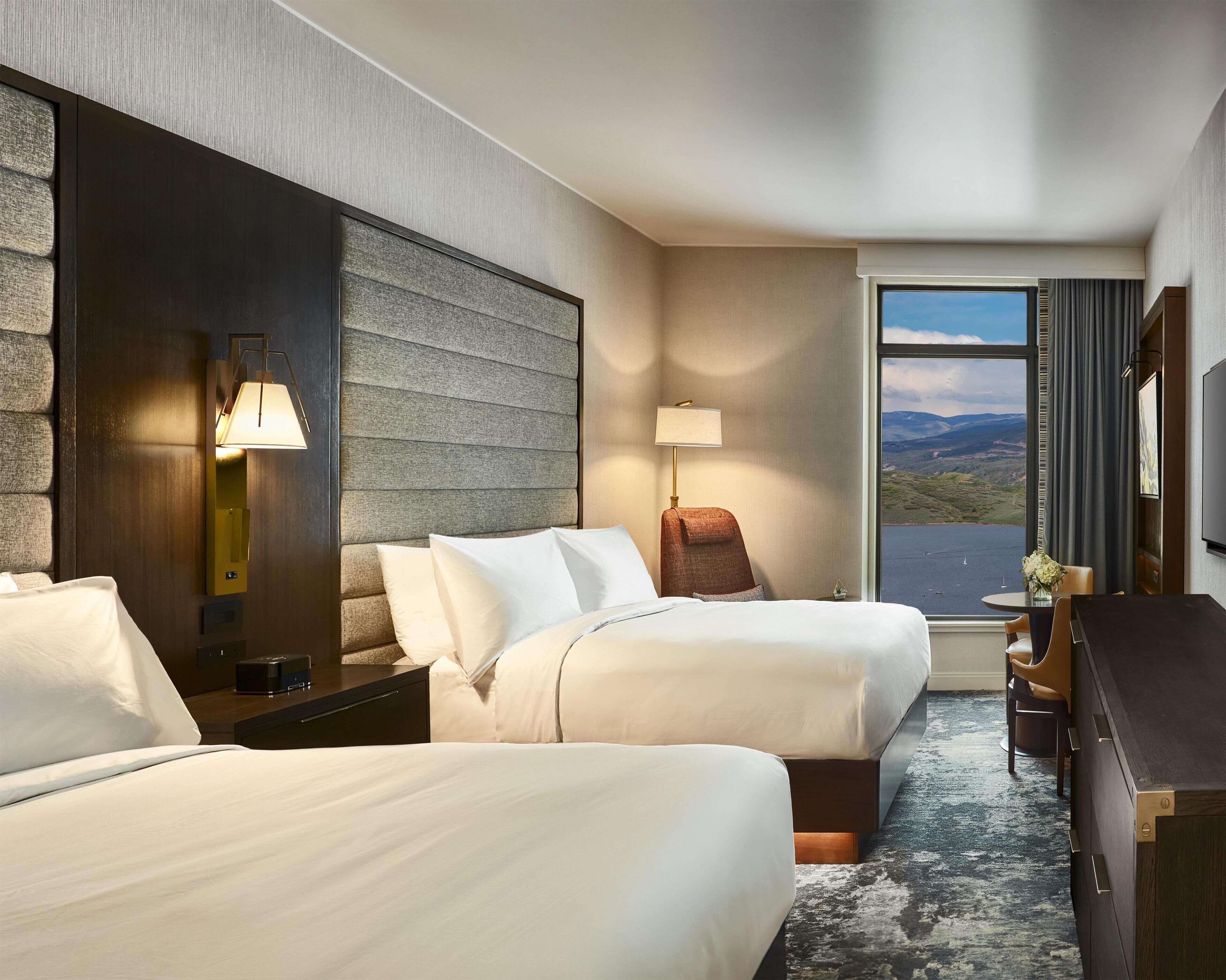 A hotel room with two beds, a desk, a chair, a lamp, a TV, and a large window with a view.