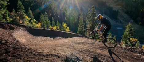 A mountain biker on a dirt trail with a forested backdrop and a clear sky.
