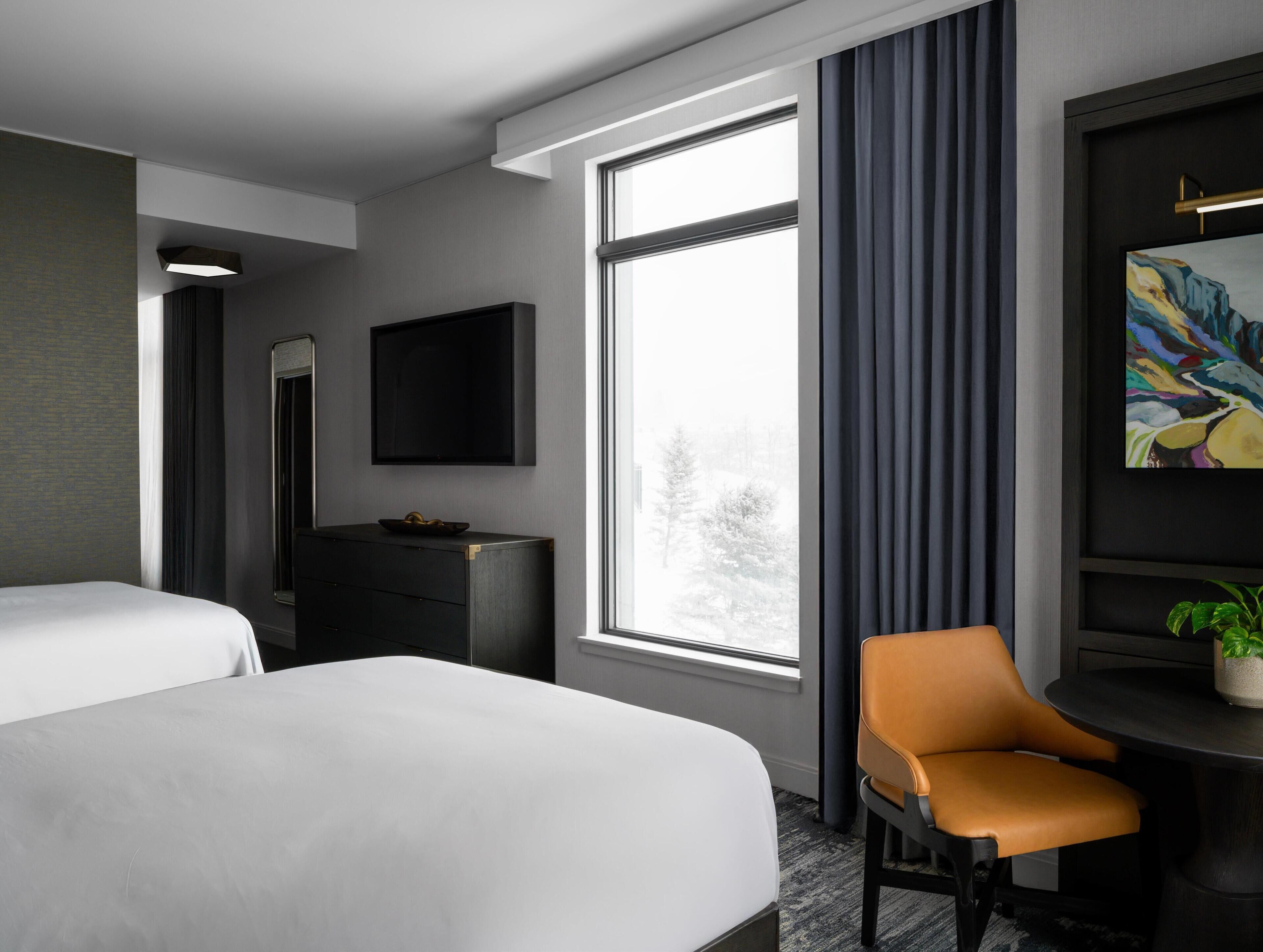 A hotel room with two beds, a desk, a chair, a TV, and a large window.