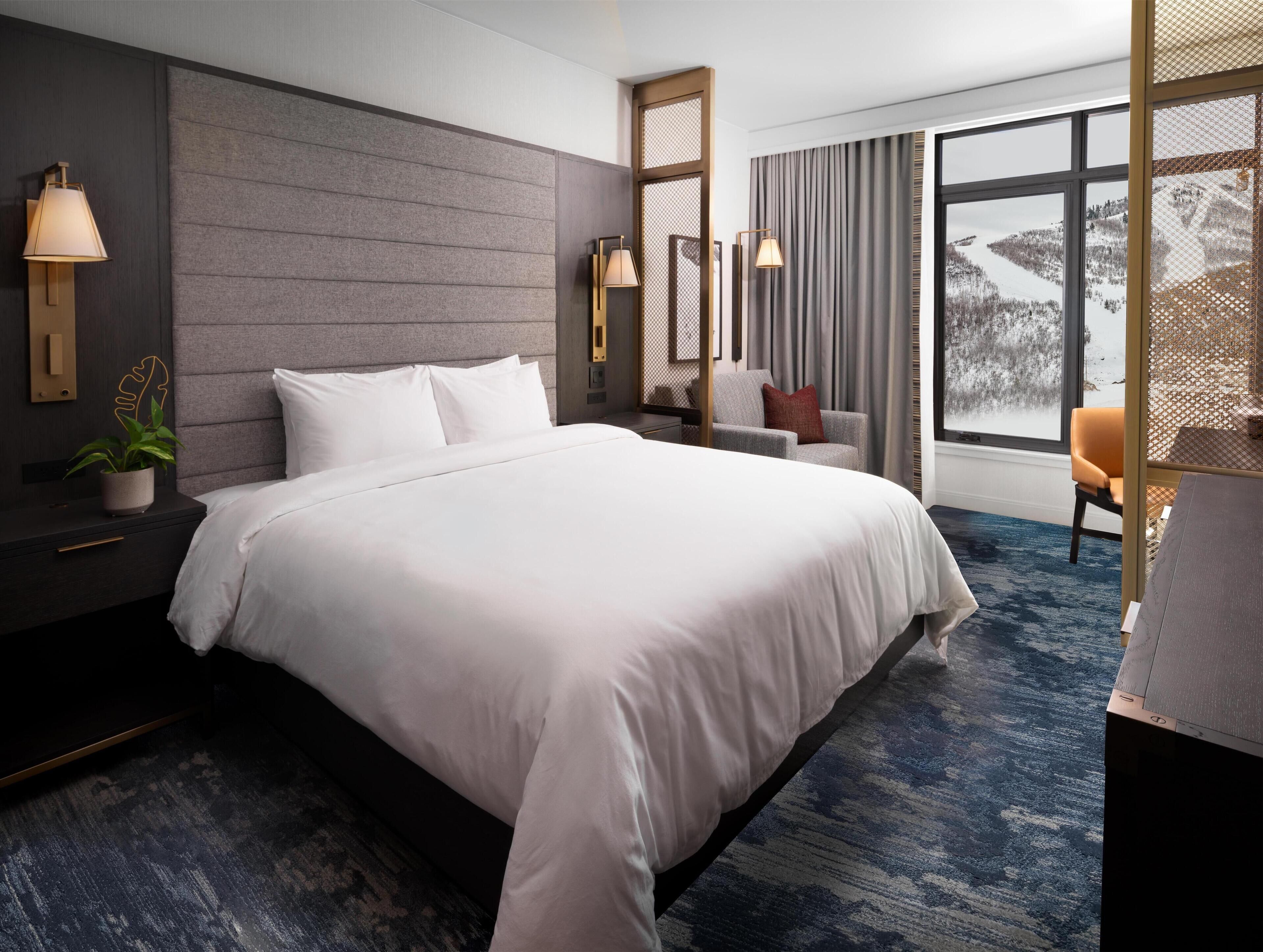 A modern hotel room with a large bed, bedside lamps, a chair, and a view of a snowy landscape.