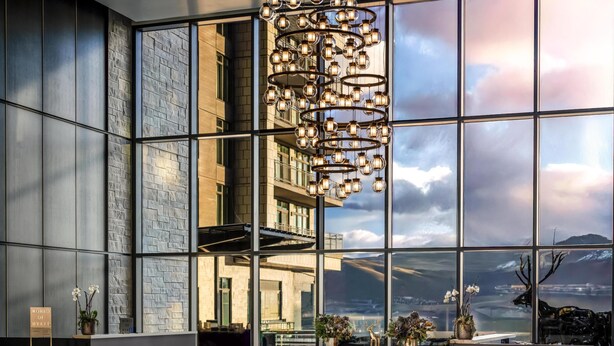 Um hall de hotel moderno com grandes janelas, um candelabro e uma mesa de recepção.