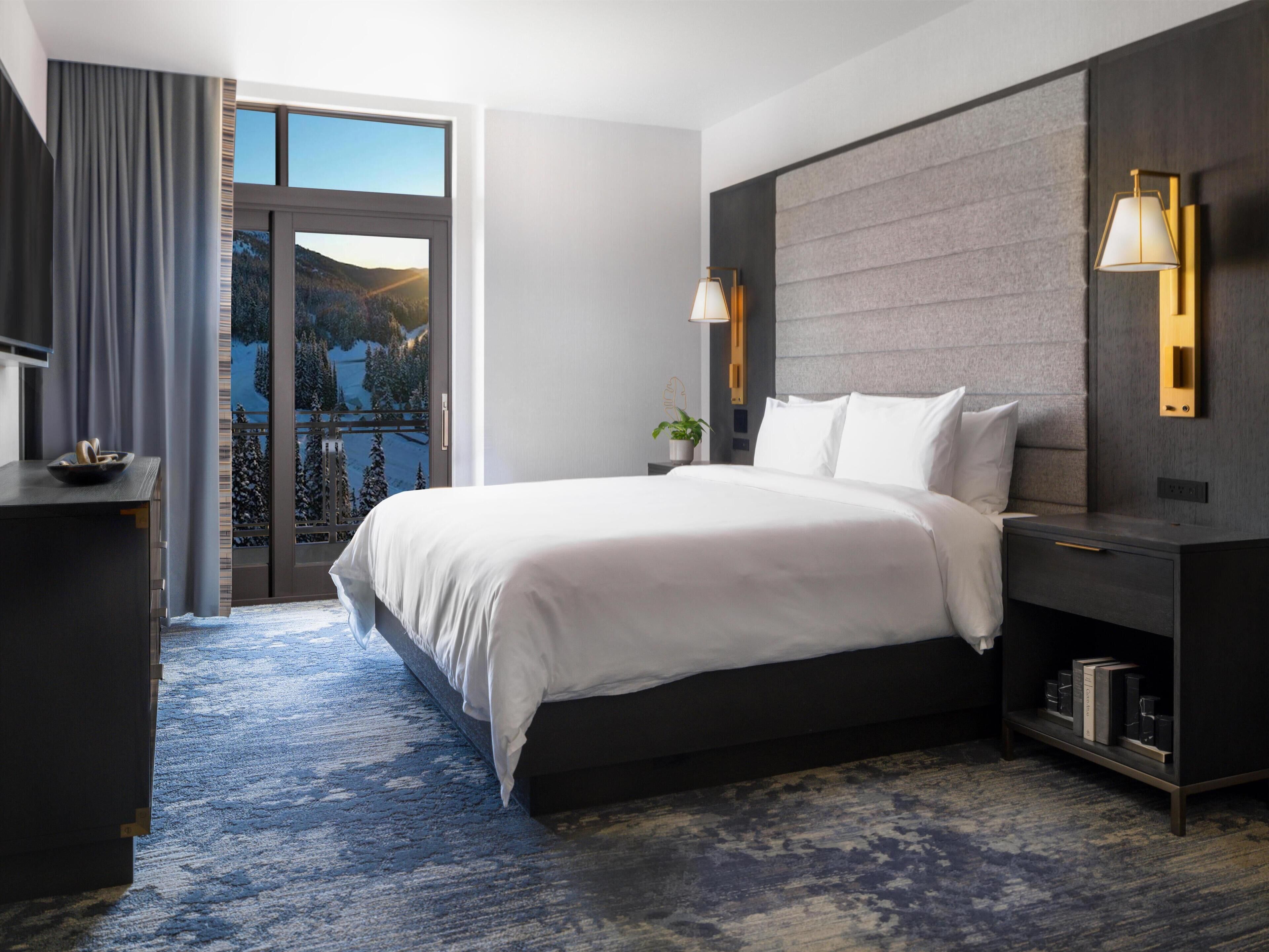 Une chambre d’hôtel moderne, dotée d’un grand lit, d’un bureau, d’une table de chevet, d’une lampe et offrant une vue sur un paysage enneigé.