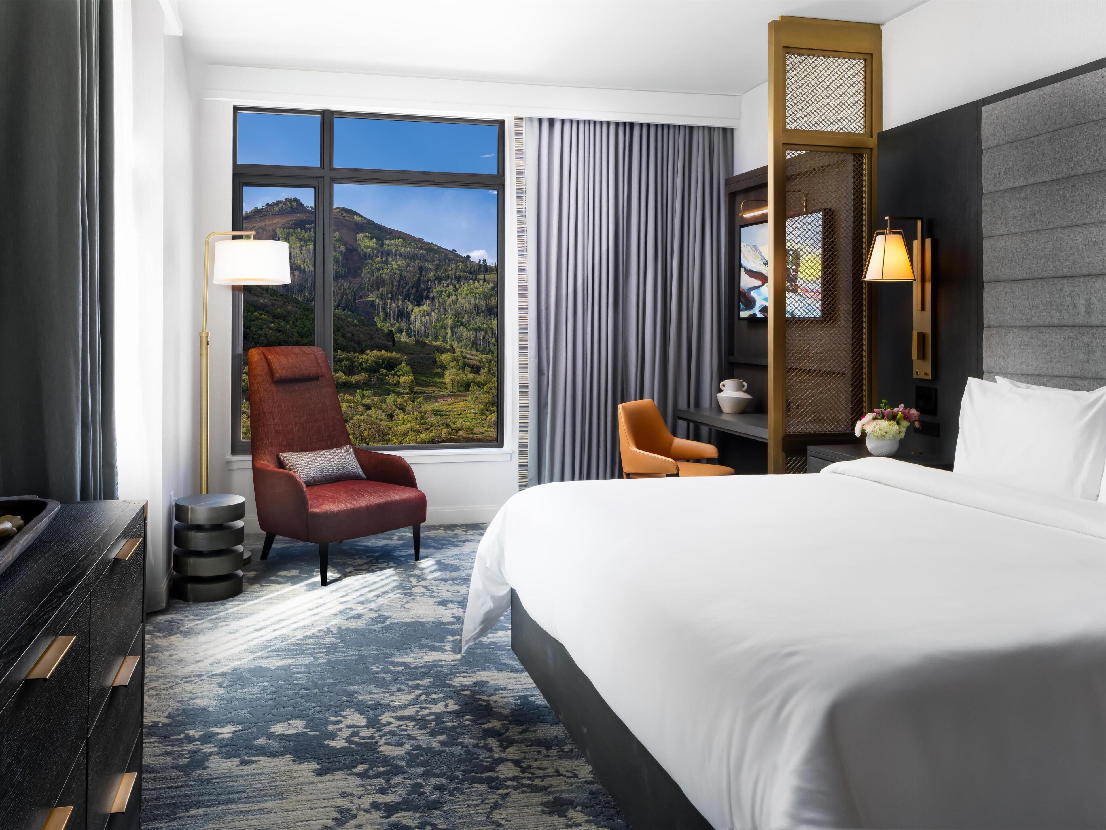 Habitación de hotel con una cama grande, dos sillas, un escritorio, una lámpara, un jarrón con flores y vistas a un paisaje montañoso.