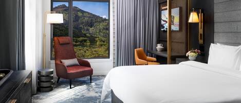 Habitación de hotel con una cama grande, dos sillas, un escritorio, una lámpara, un jarrón con flores y vistas a un paisaje montañoso.