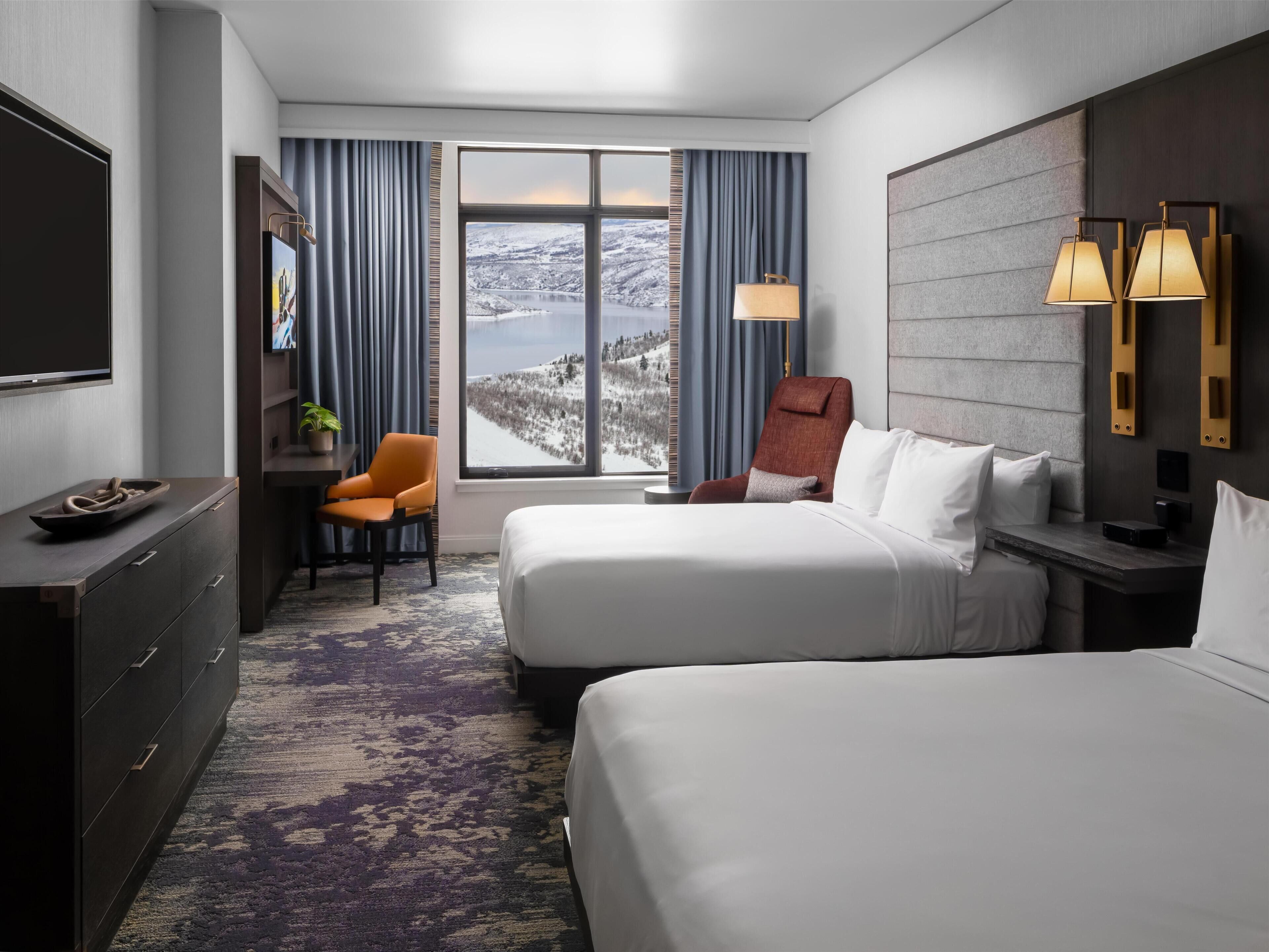 Ein Hotelzimmer mit zwei Betten, einem Schreibtisch, einem Stuhl, einem Fernseher, einem Fenster mit Blick auf eine verschneite Landschaft und zwei Wandleuchten.