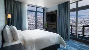 Quarto de hotel com uma cama grande, televisão, secretária e vista para uma paisagem nevada.