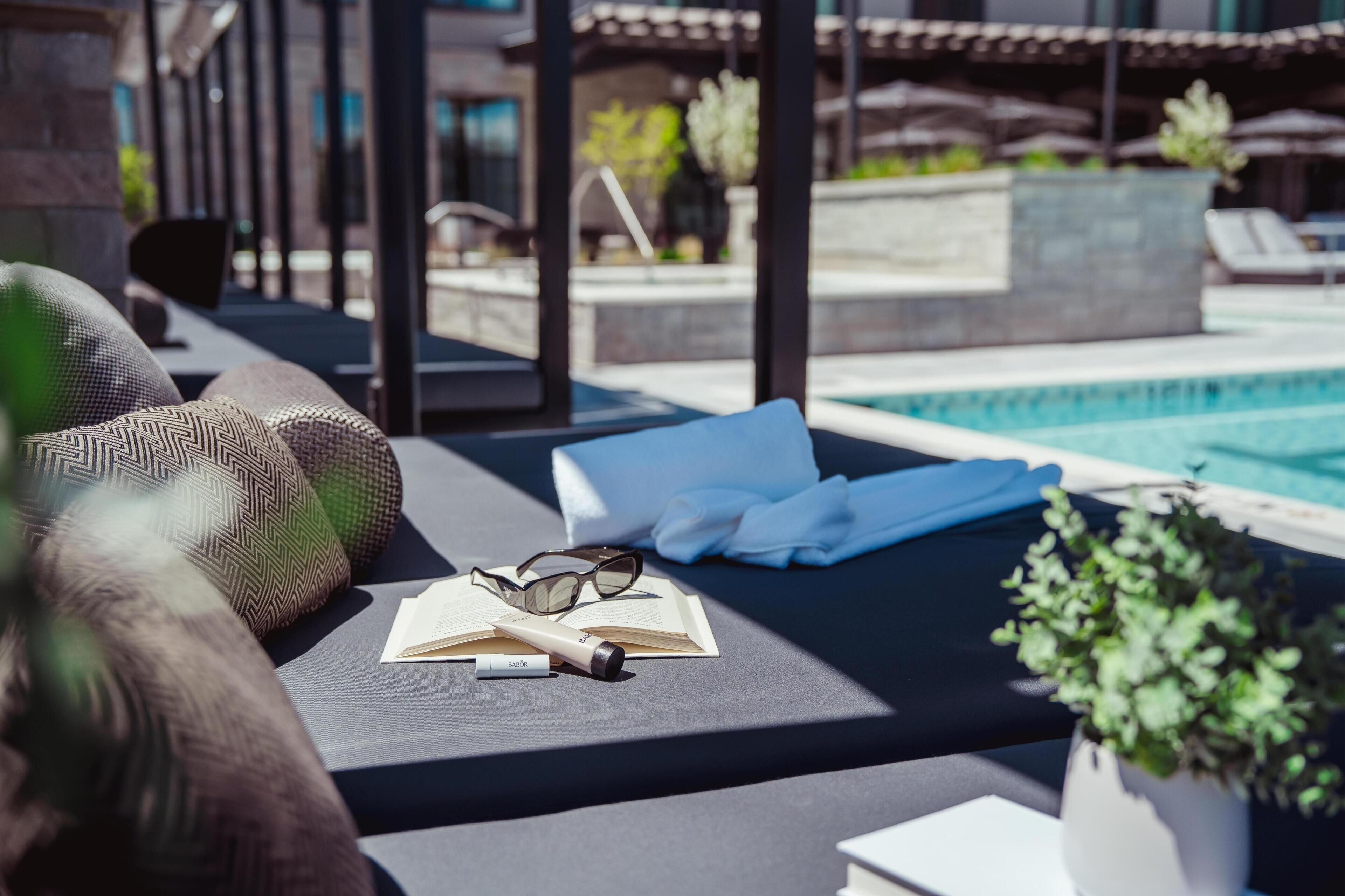 Zona de descanso junto a la piscina con un sofá, un par de gafas, un libro y una botella, con piscina y sombrillas al fondo.