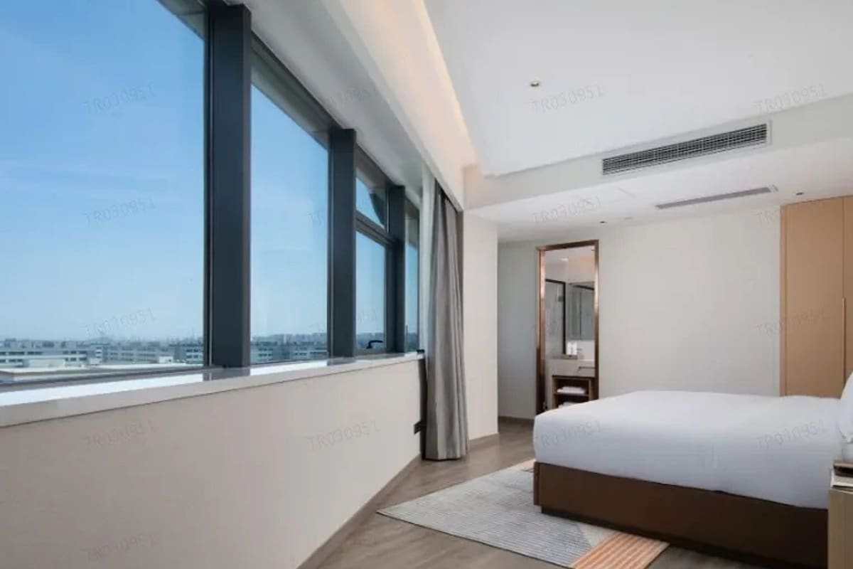 A modern hotel room with a large window, a bed, a nightstand, and a mirror.