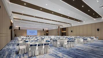 A large conference room with rows of round tables and white chairs, a large screen, and a blue carpeted floor.
