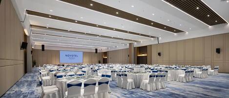 A large conference room with rows of round tables and white chairs, a large screen, and a blue carpeted floor.