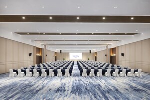 Un salón de conferencias con filas de sillas y mesas, una pantalla grande al frente y un diseño de techo moderno.