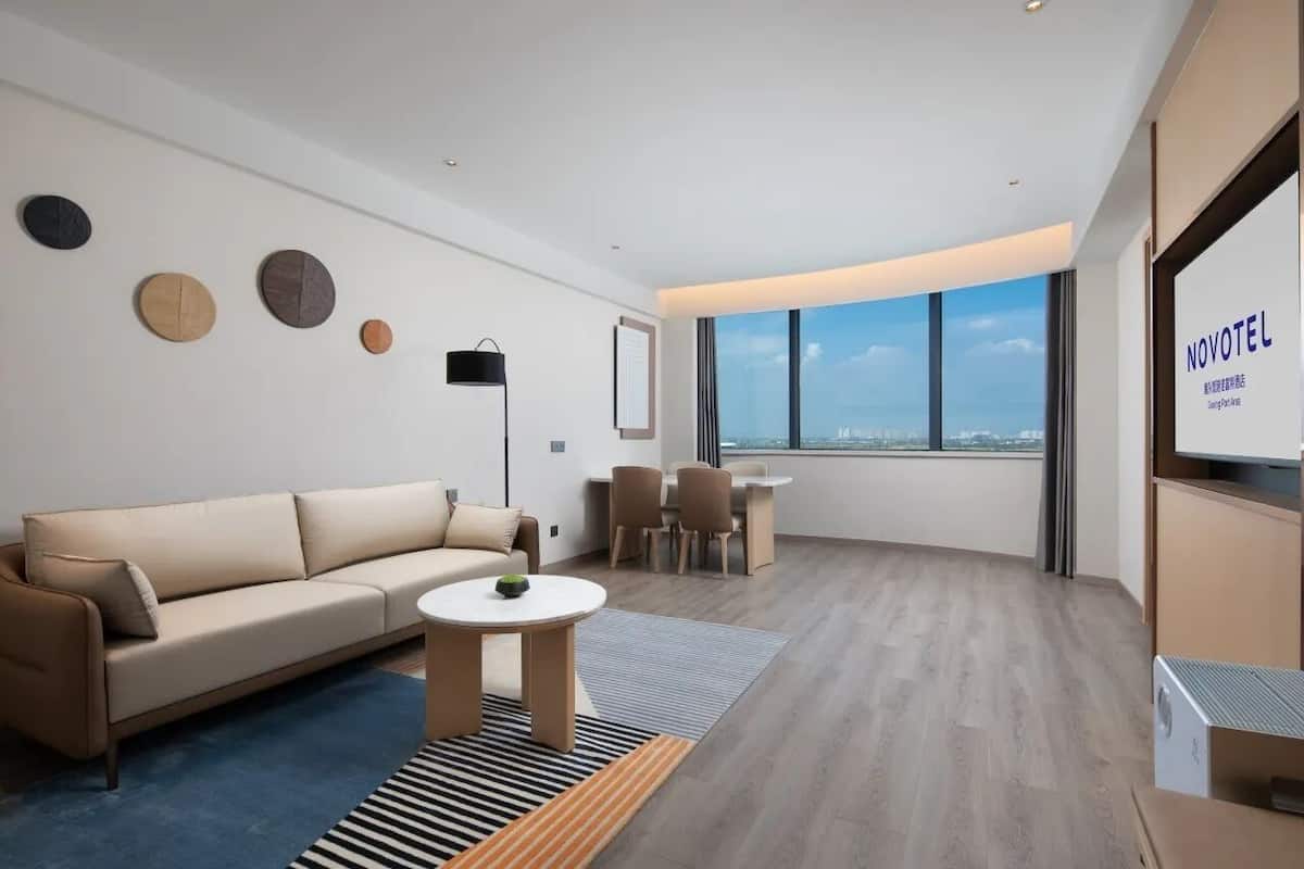 A modern hotel room with a sofa, a small round table, and a view of the city through large windows.